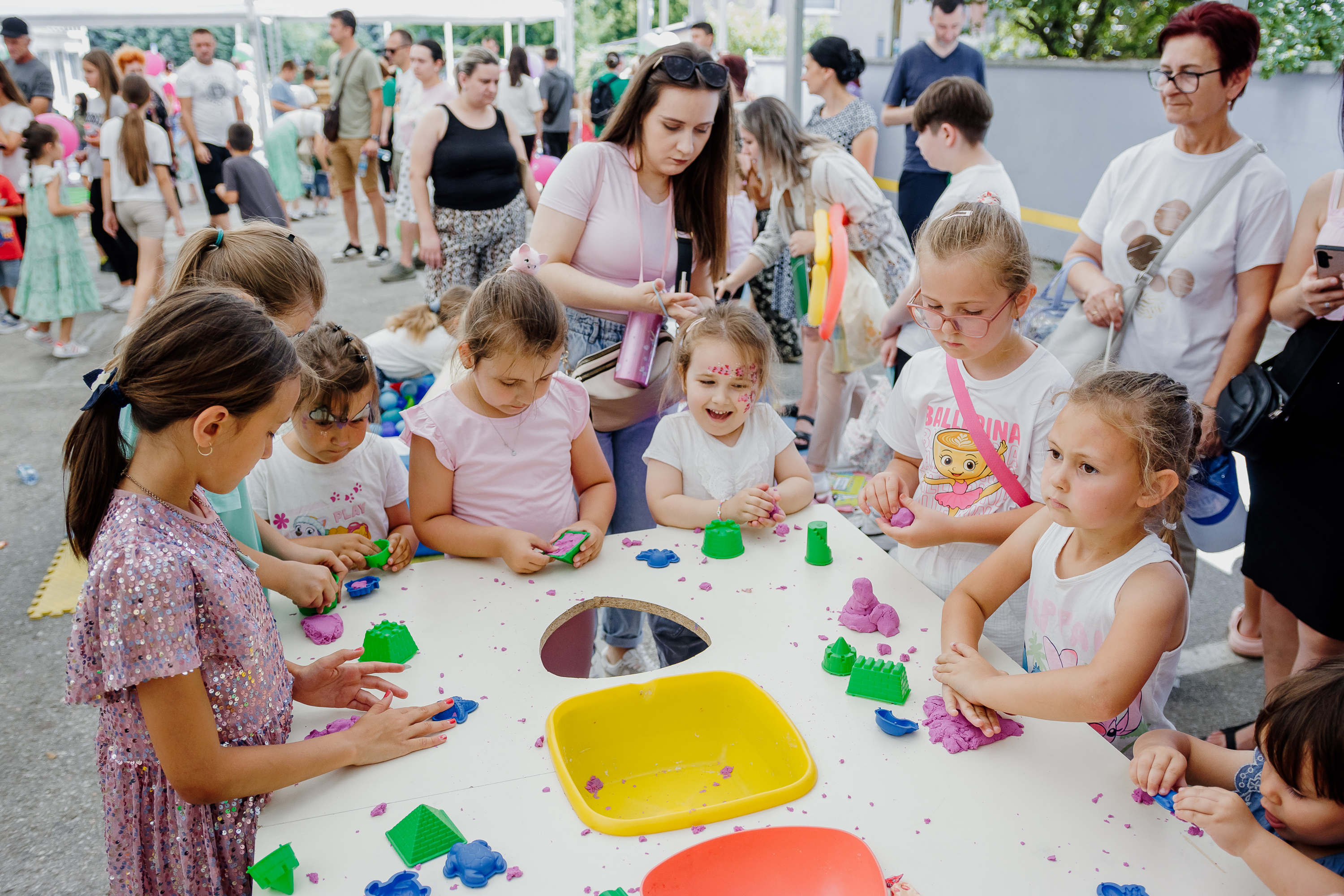 Djeca sjede za velikim okruglim stolom na otvorenom i igraju se oblicima od plastelina u različitim bojama. Na stolu su razbacani šareni komadi plastelina, kalupi i posude. Odrasli stoje u pozadini, posmatrajući aktivnost.