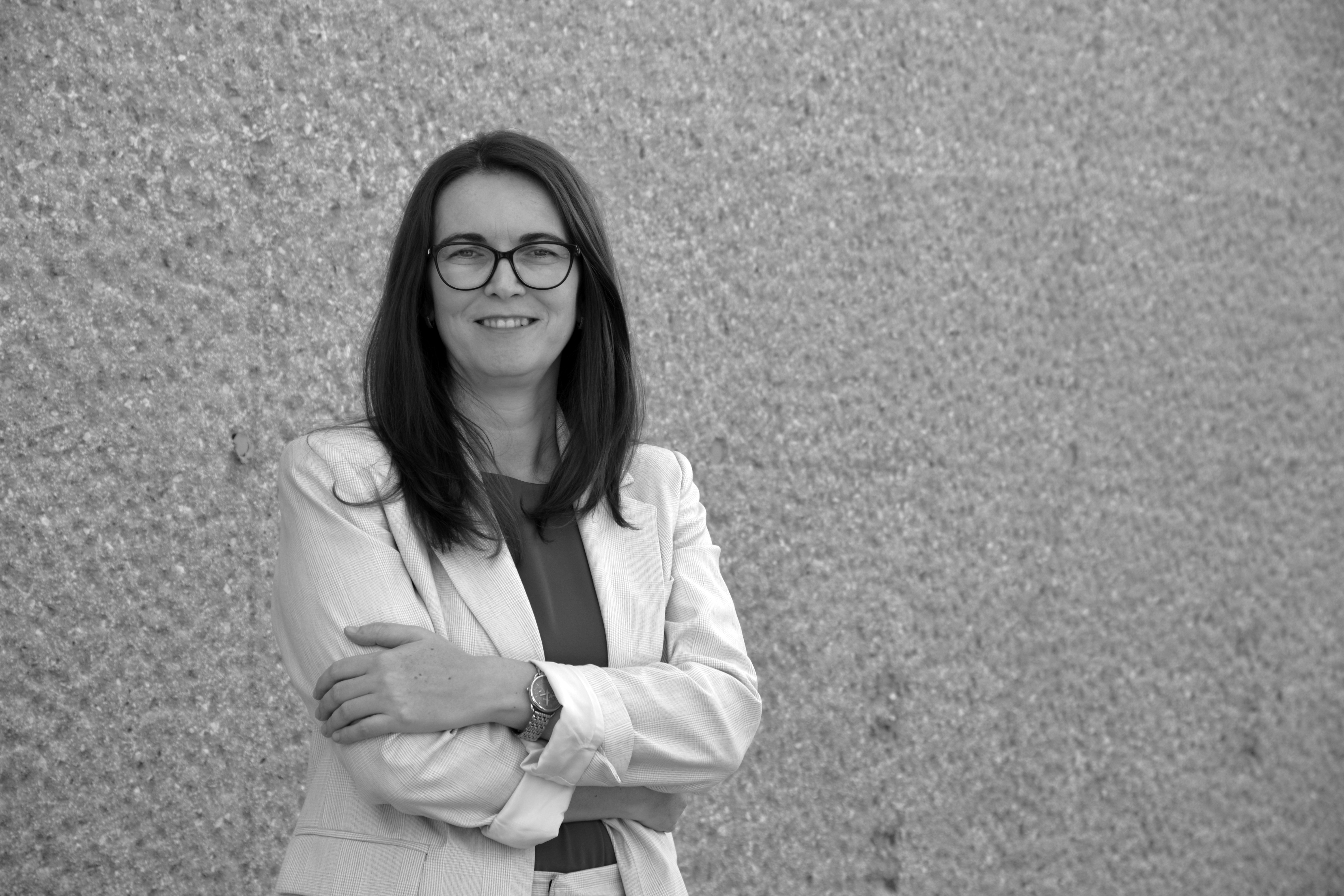 Žena s dugom kosom stoji ispred grube teksturirane zidne pozadine, nosi bijeli sako s prekriženim rukama.