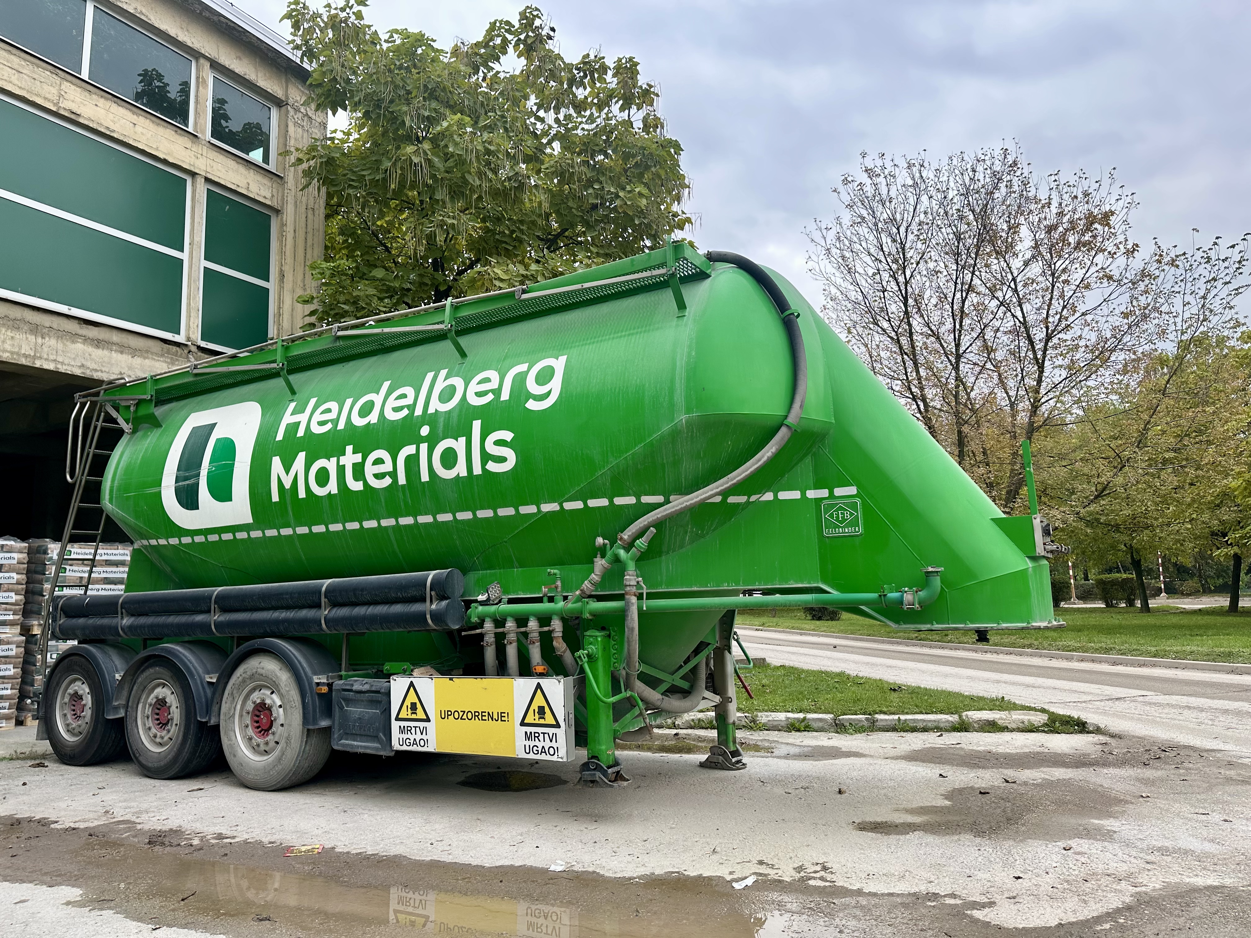 Zeleni kamion-cisterna sa logotipom "Heidelberg Materials" parkiran ispred poslovne zgrade, okružen drvećem i betonskim putem.
