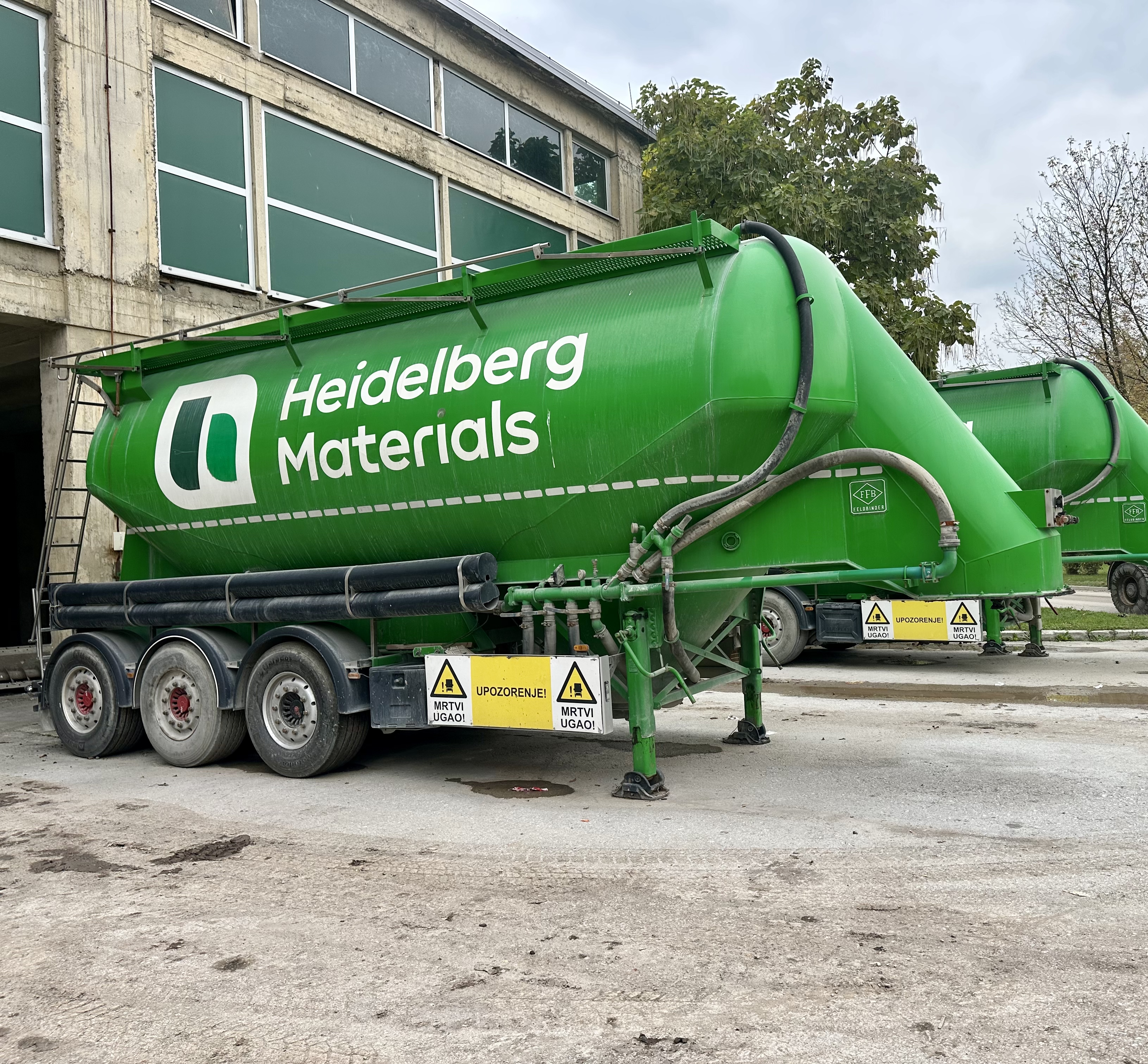 Zeleni kamioni-cisterne s logotipom Heidelberg Materials parkirani ispred industrijske zgrade, s drvećem u pozadini.