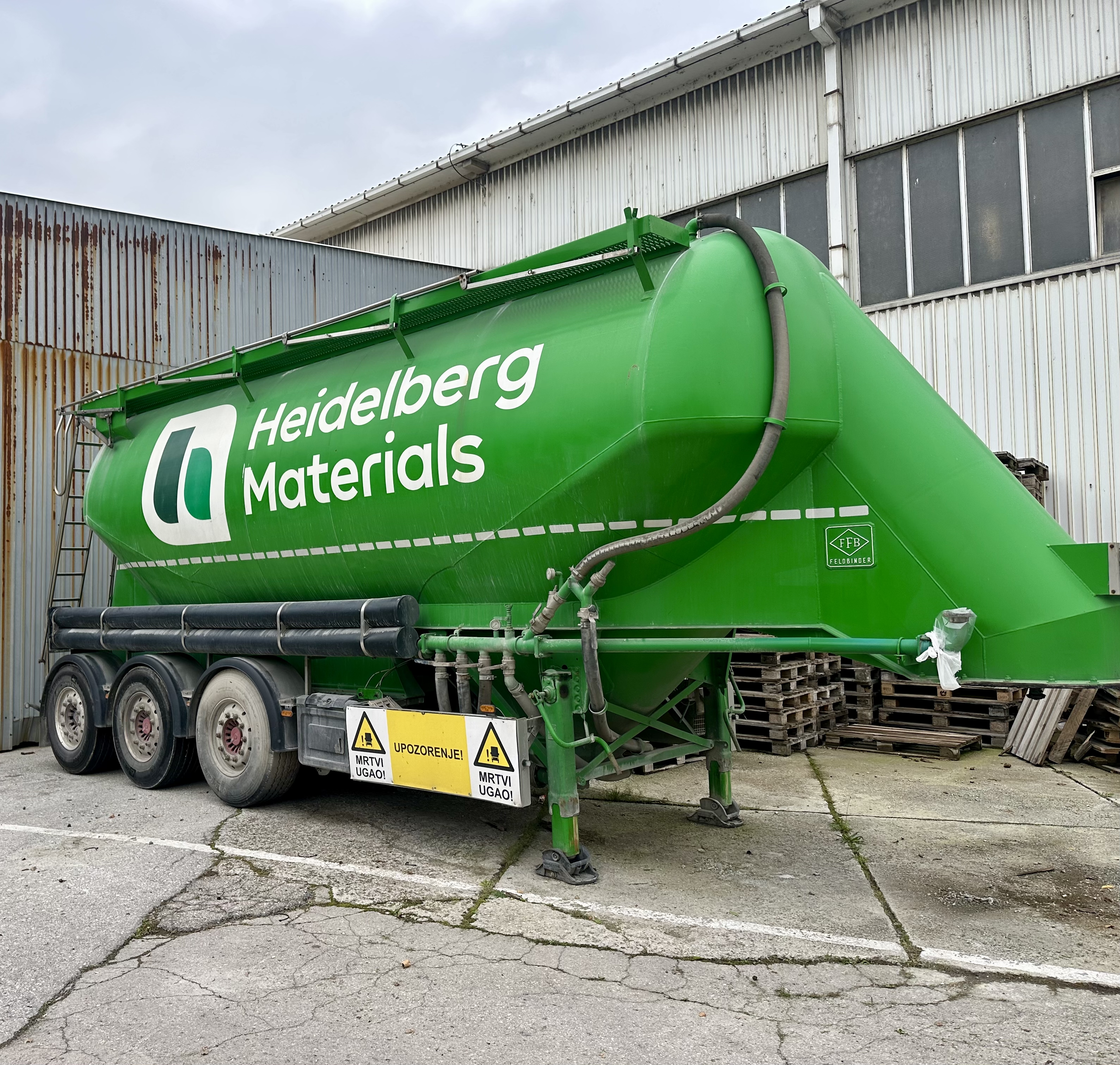 Zeleni cistercijski prikolica s natpisom "Heidelberg Materials" parkirana ispred industrijskog objekta, sa sigurnosnim oznakama na donjem dijelu prikolice i sivim metalnim zidovima u pozadini.