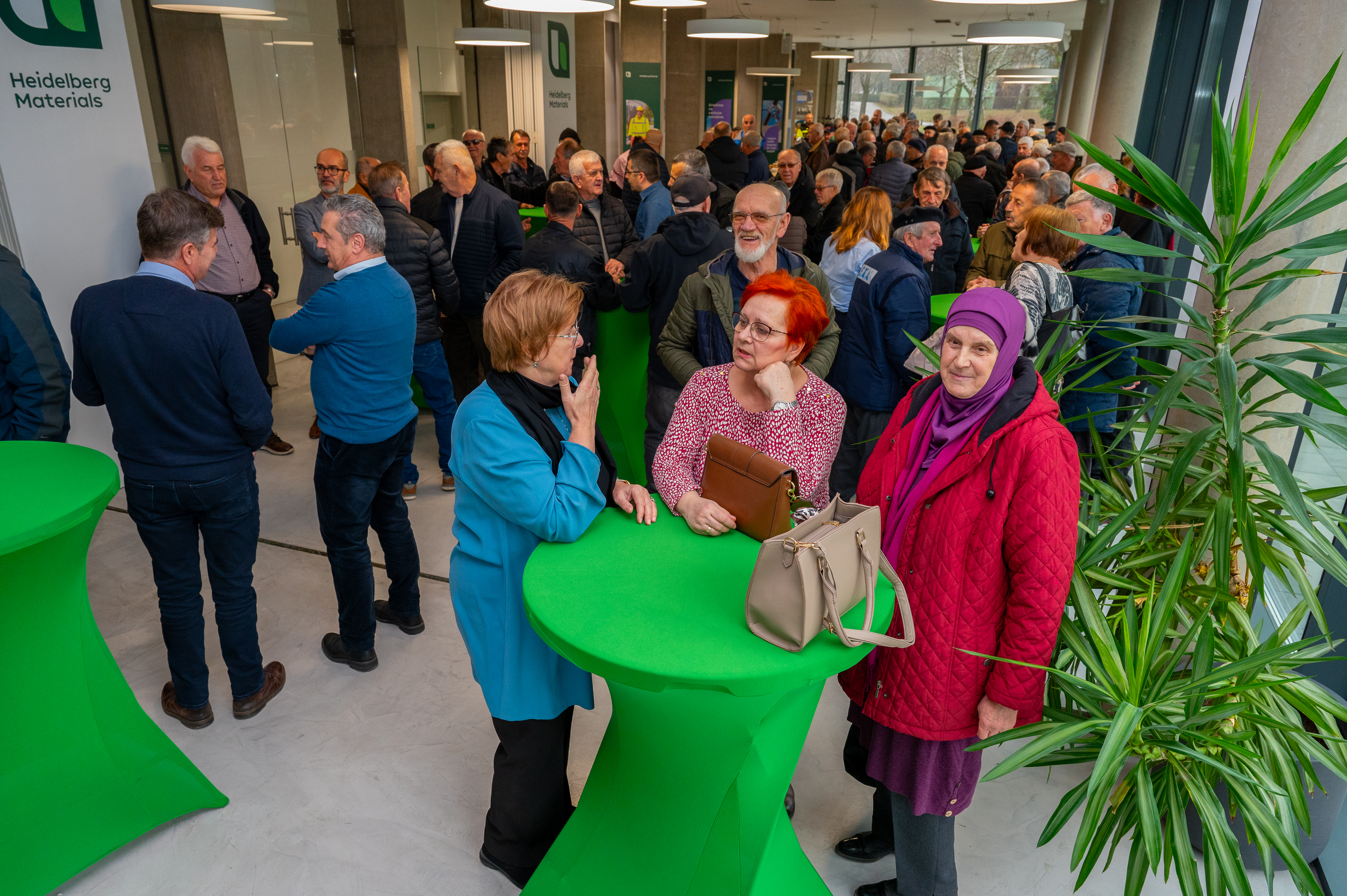 Grupa ljudi okupljena u moderno uređenom prostoru sa zelenim stolovima, razgovaraju u manjoj i većoj grupi. U prvom planu su tri žene kod jednog stola, okružene ostalim učesnicima događaja. Na desnoj strani nalazi se velika zelena biljka.