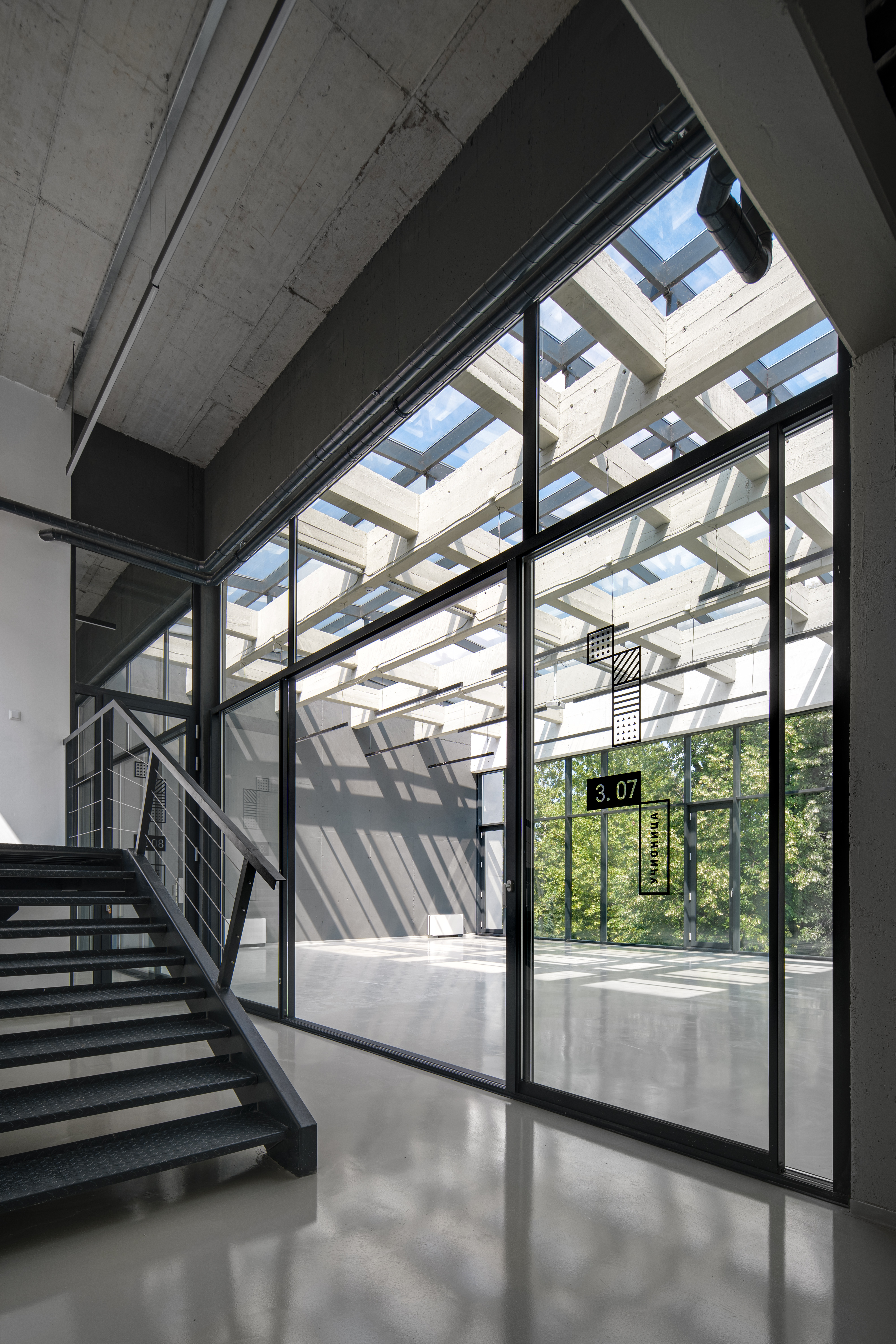 Unutrašnjost moderne zgrade sa staklenim zidovima i velikim prozorima, kroz koje dopire prirodno svjetlo. Lijevo se nalazi metalno stepenište, dok se kroz prozore vidi zelenilo na otvorenom. Pod je svijetao i reflektivan, a strop visok s vidljivim konstrukcijama.