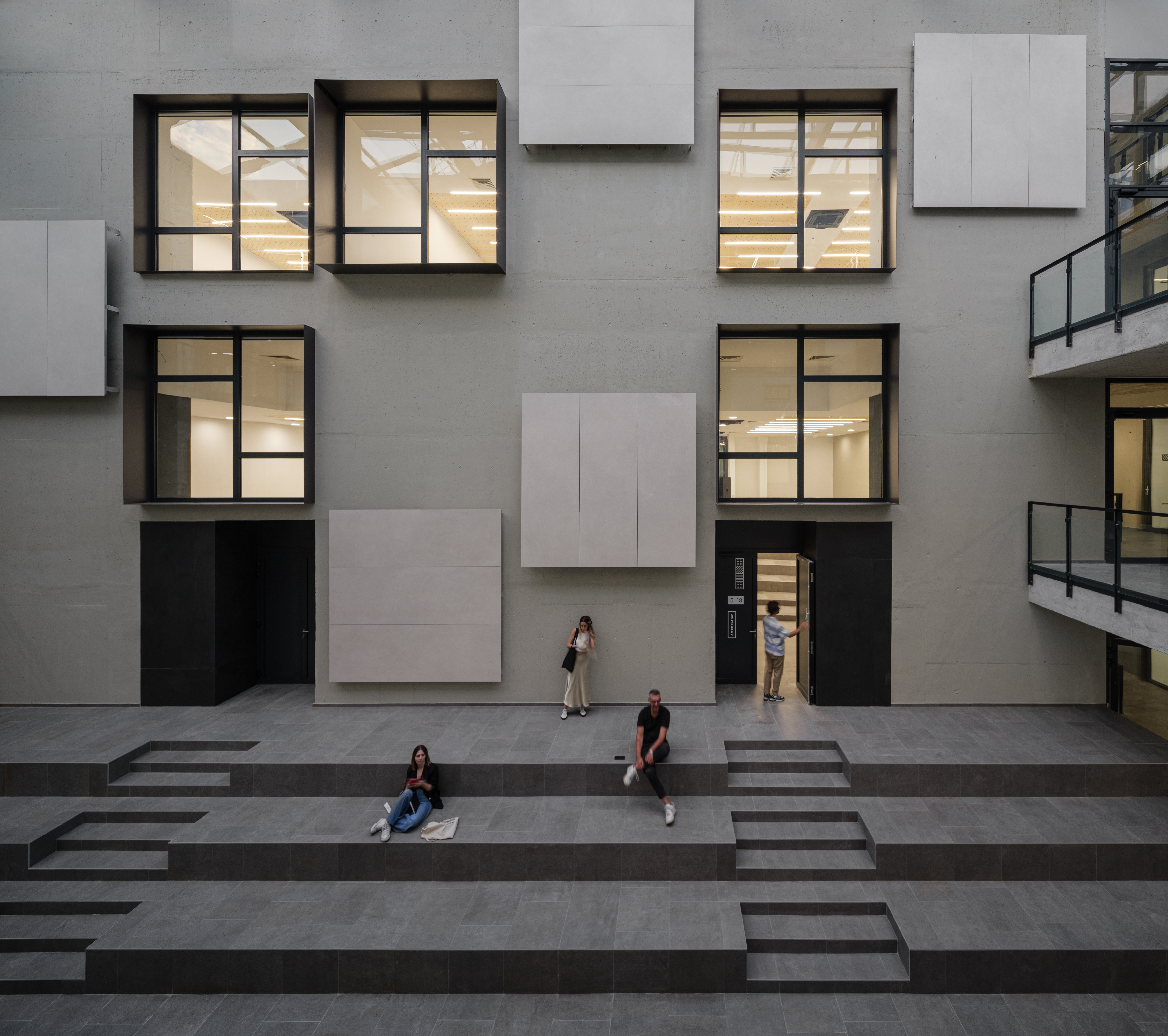 Fasada moderne zgrade s velikim prozorima i geometrijskim panelima. Ispred zgrade nalaze se stepenaste klupe na kojima sjede tri osobe dok jedna osoba stoji ispred ulaza. Unutrašnjost zgrade osvijetljena je prirodnim svjetlom.