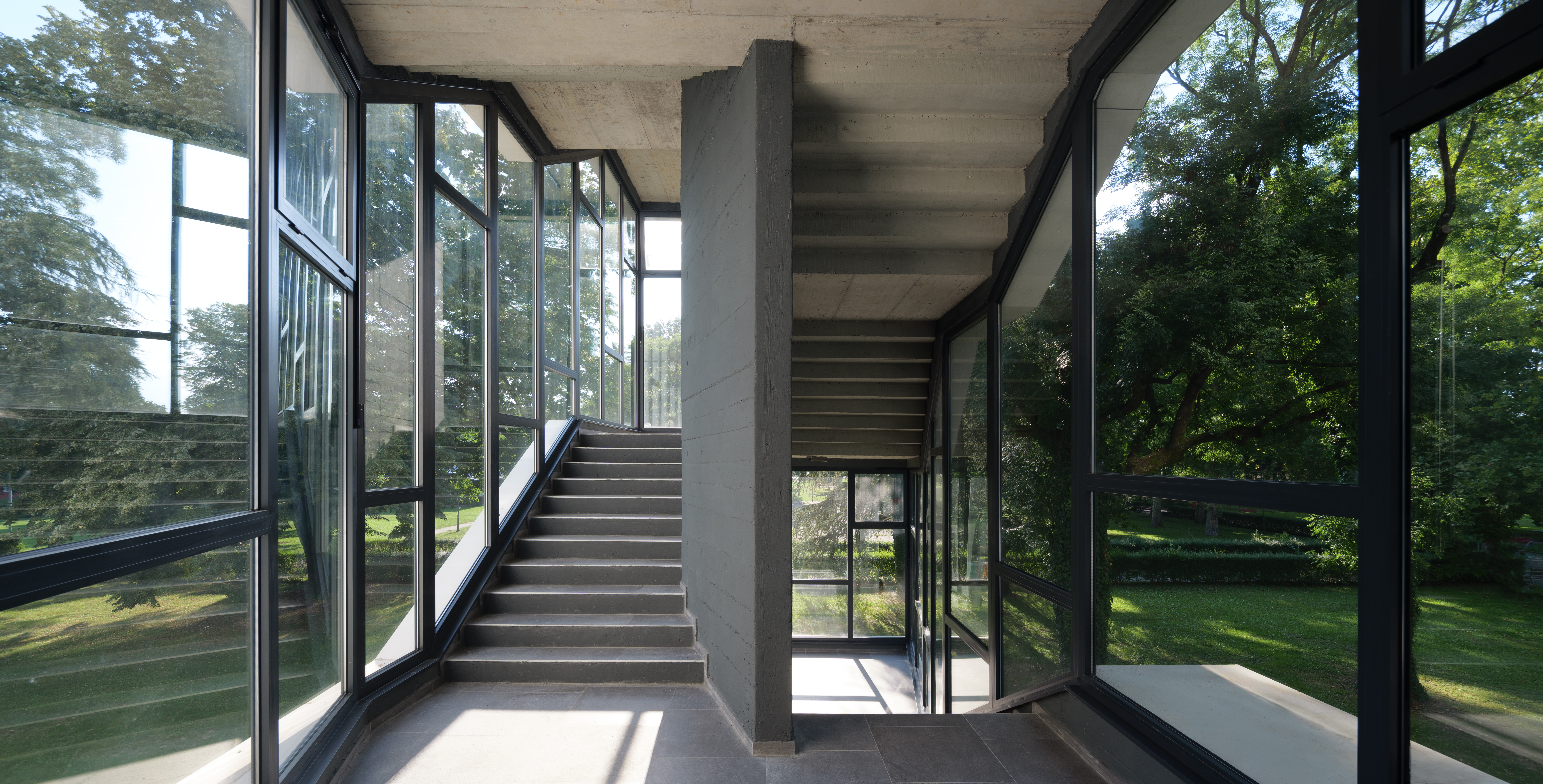 Stakleno stubište modernog dizajna s velikim prozorima na obje strane, pružajući pogled na zelenu okolinu. Stubište se proteže naviše, dok se prirodna svjetlost reflektira kroz prozore, naglašavajući minimalističku arhitekturu.
