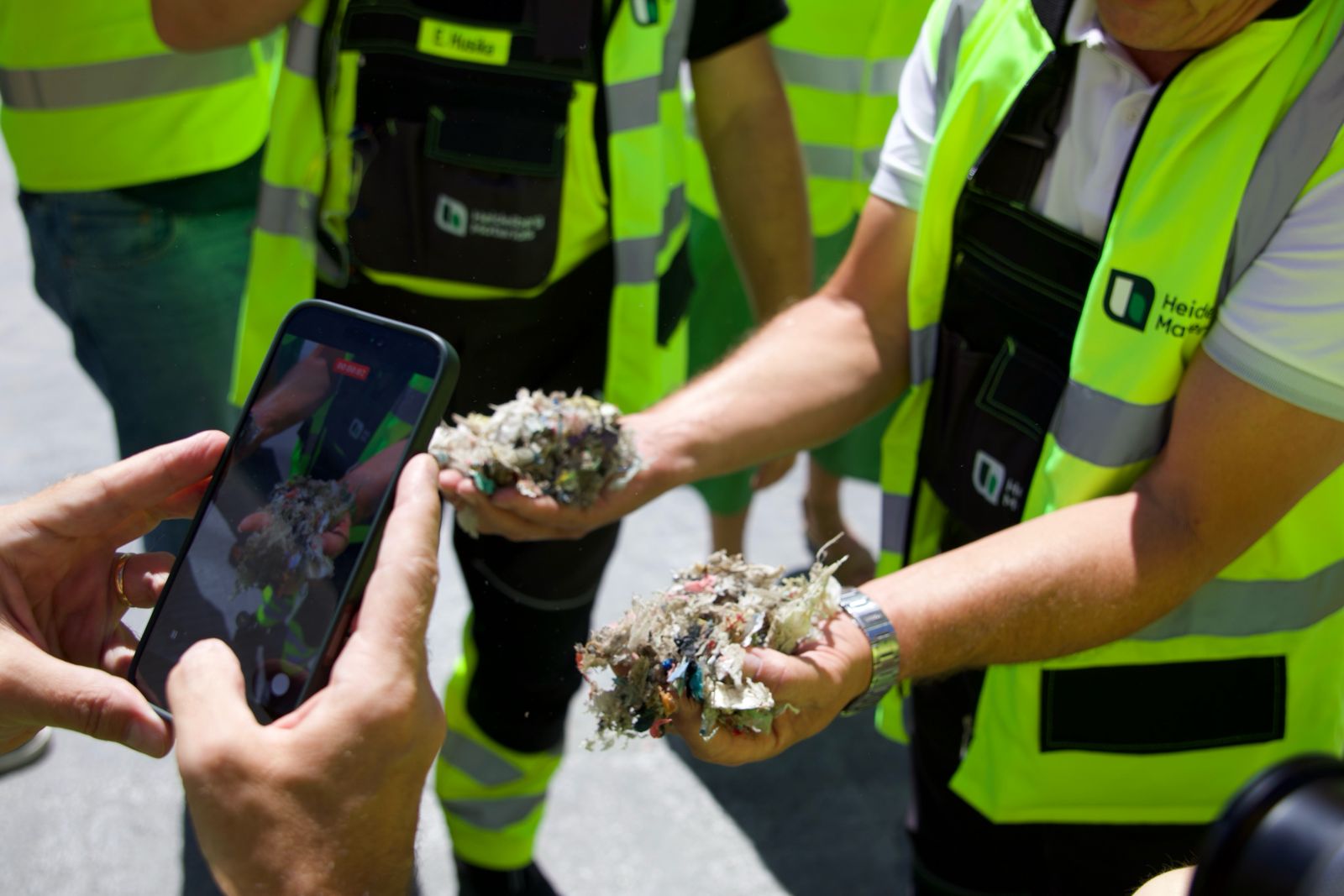 Osoba u reflektirajućem prsluku drži šaku drobljenog materijala dok druga osoba fotografiše mobilnim telefonom.