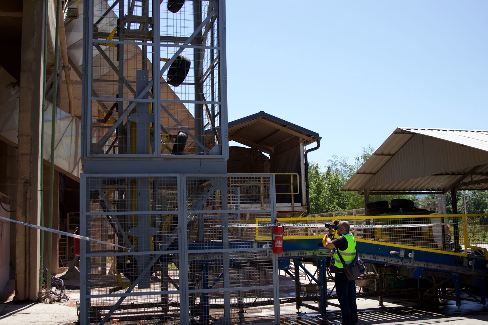 Radnik u sigurnosnom prsluku stoji pored industrijskog postrojenja na otvorenom.