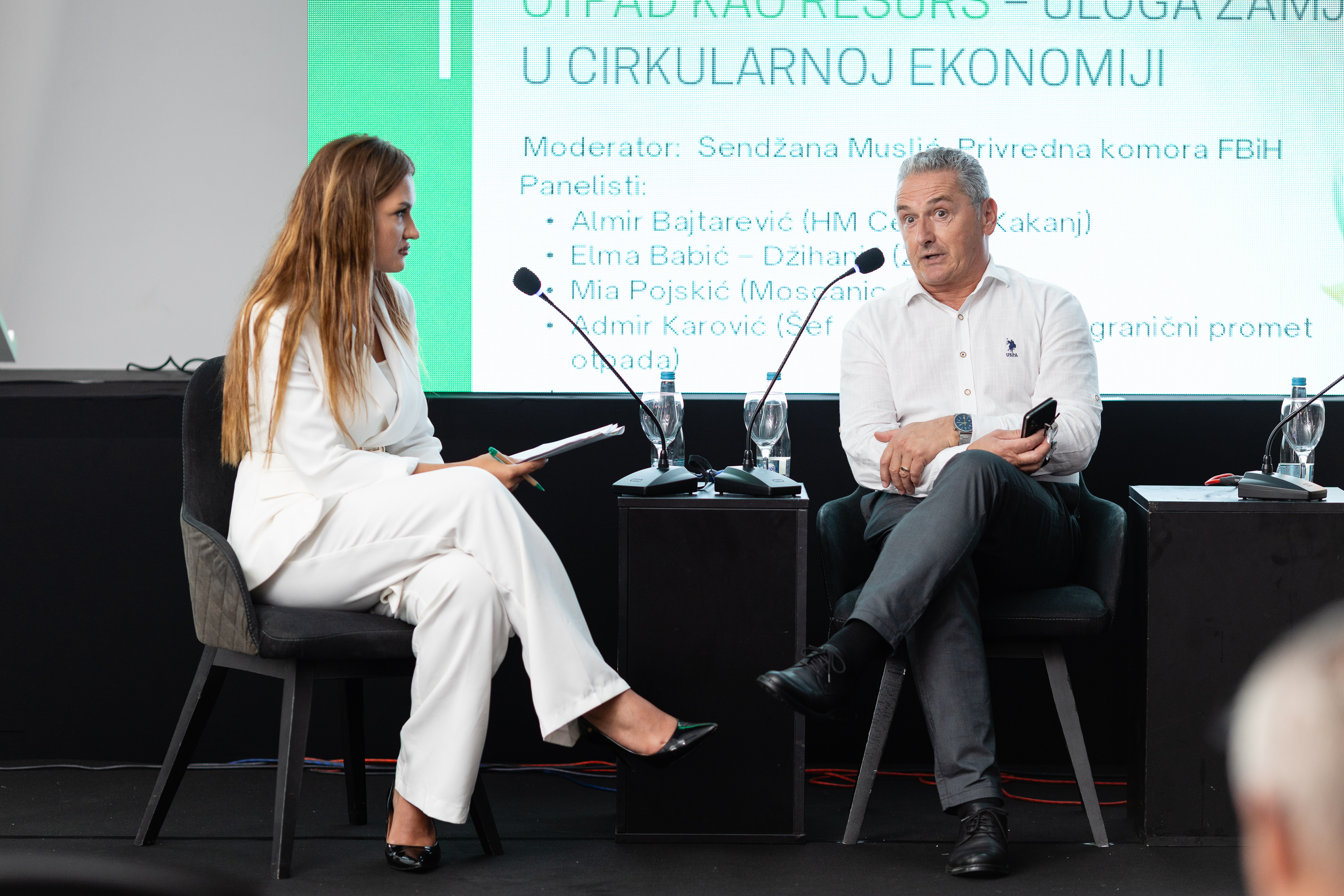 Dvoje ljudi razgovaraju na panelu o cirkularnoj ekonomiji, uz projekciju teme događaja na ekran u pozadini.