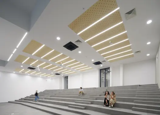 Amfiteatar s modernim dizajnom, stepenastim sjedištima od betona i nekoliko osoba koje sjede razmaknuto. Plafon ima drvene akustične panele i ugradbena LED svjetla. U pozadini su staklena vrata. Prostor je svijetao, minimalistički uređen.